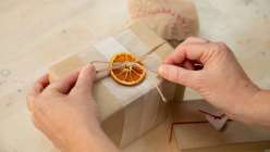 A pair of women's hands tying a piece of twine around a brown-paper package, looped through a dried slice of orange.