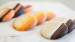 Three sets of three sugar cookies dipped into ombre-effect icing, one with an orange to black gradient, one with a yellow to brown gradient, and one with a white to black gradient. 