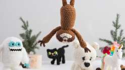 An adorable crocheted amigurumi sloth hanging upside-down in front an assortment of crochet friends, including a crochet yeti, a crochet black cat, and a crochet polar bear.